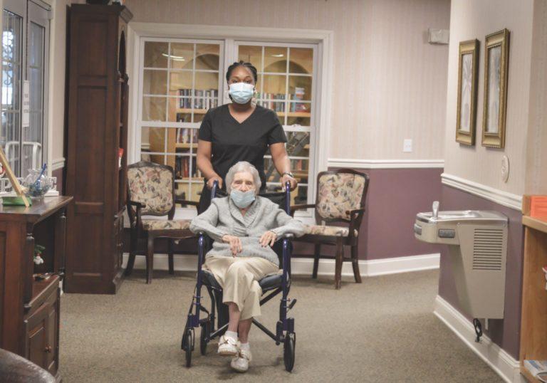 caregiver in facemask pushing assisted living resident with facemask in wheel chair