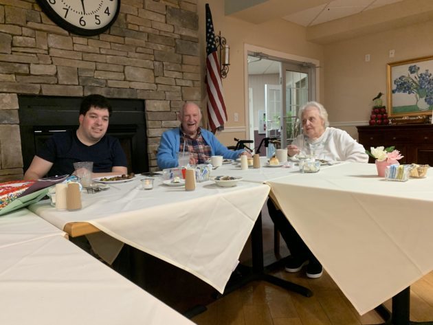 smiling man and woman residents sitting at dining table enjoying meal with caregiver